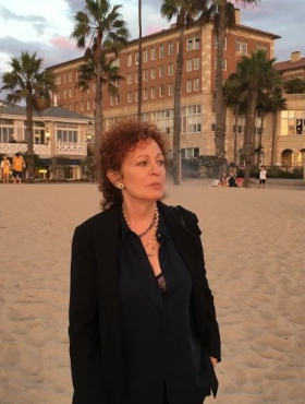 Nan Goldin steht an einem Strand und betrachtet die Umgebung. Im Hintergrund sind einige Gebäude, Palmen und Personen zu sehen.